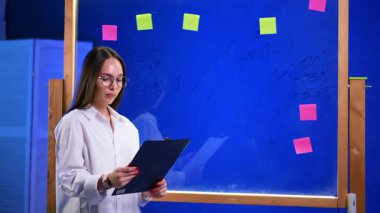 Gözlüklü beyaz bir kadın elindeki panoya bakmış. Kızın arkasındaki cam duvarda çıkartma notları ve yazılı numaralar var..