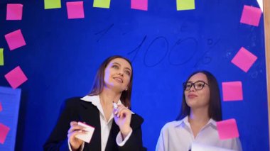 Beyaz kadınlar cam duvarın arkasında dururlar. Ceketli kadın meslektaşıyla konuşur ve tahtaya yazı yazar..