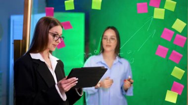 Cam duvarın iki tarafında da iki beyaz kadın var. Önde duran kadın tableti tutuyor. Tahtanın arkasındaki kız bir kalemle yazıyor..