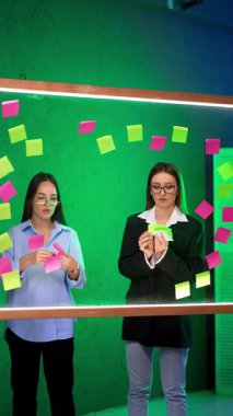 Cam duvarın arkasında duran iki odaklı bayan. Notlar yazıp planlama için tahtaya koyuyorum. Dikey video.