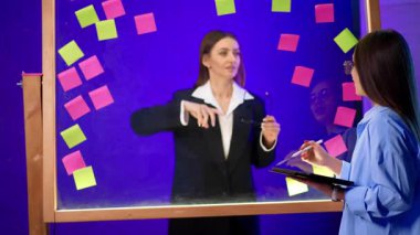 Kadınlar cam duvara bakar, konuşur ve çıkartmaları tartışırlar. Öndeki kız tableti gösteriyor.