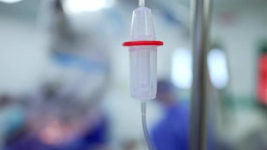 Akciğer havalandırma makinesinin yanındaki kürsüde asılı duran konteynır. Cerrahi ekip bulanık arkaplanda ameliyatta çalışıyor..