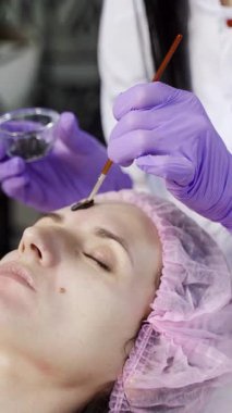 Güzellik uzmanı bir güzellik salonunda müşterinin yüzüne siyah bir maske takar. Cilt bakımı. Güzellik endüstrisi Dikey video