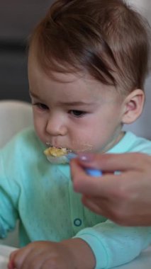 Metal kaşıkla oynayan sevimli bir bebek annemden beslenir. Mavi gömlekli pis çocuk beyaz masada oturup kaşıkla yemeğini yiyor. Arkaplan bulanık. Dikey video