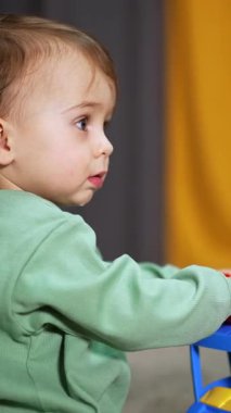 Açık yeşil tişörtlü küçük bebek elinde büyük renkli bir kamyon tutuyor. Kahverengi saçlı çocuk odada oyuncakla oynuyor ve etrafa bakıyor. Arkaplan bulanık. Dikey video