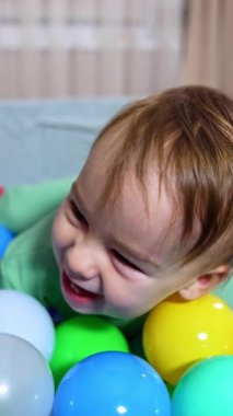 Mutlu erkek bebek evde heyecanlı bir şekilde oynuyor. Küçük beyaz çocuk yumuşak havzadaki renkli topların arasında uzanıyor. Arkaplan bulanık. Dikey video