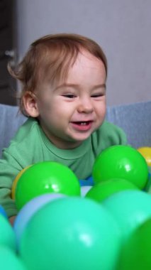 Bir yaşındaki bir çocuk küre ve toplarla havuzda oynuyor. Tatlı çocuk oyuncaklara atlarken mutlu bir şekilde gülümsüyor. Kapatın. Arkaplan bulanık. Dikey video