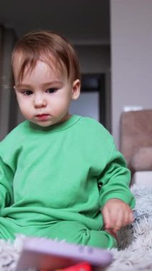 Tatlı küçük bebek, odadaki yumuşak yumuşak bir halıya oturur. Bebek telefonda video izleyip el çırpıyor. Düşük açı. Dikey video