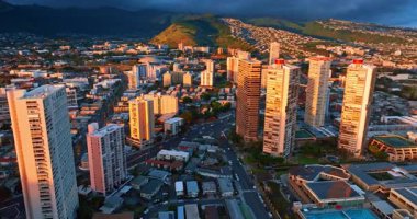 Gün batımının turuncu ışığında Honolulu 'nun gökdelenleri. Şehirden geçen yol boyunca birden fazla araba hareket ediyor. Yeşil dağlar arka planda kara bulutlarla kaplıdır..