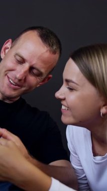 Babası küçük, ciddi bebeğini kucağına almış. Baba çocuğun elini sallıyor ve anne oğluna gülümsüyor. Dikey video