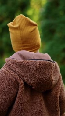 Kahverengi ceketli, sarı şapkalı ve pantolonlu bir çocuğun arka görüntüsü. Çocuk parktan kaçıyor. Dikey video