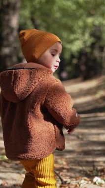 Kahverengi ceketli ve sarı şapkalı sevimli oğlanı takip ediyorum. Sonbahar parkında yürüyüş yapan tatlı çocuk. Dikey video