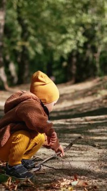 Küçük çocuk parktaki ara sokağa çömelmiş oturuyor. Çocuk sopaya dokunuyor ve ayağa kalkıyor ve uzaklaşıyor. Dikey video