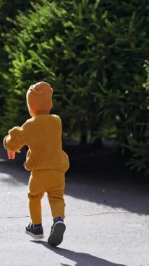 Yolda koşan mutlu çocuğu takip ediyorum. Sarı takım elbiseli ve açık havada güneşli havada şapkalı bebek. Dikey video