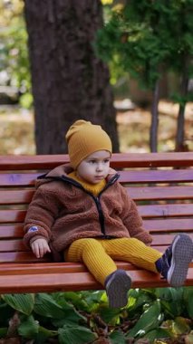 Güzel çocuk bankta oturuyor ve ayağını sallıyor. Sakin bebek parkta dinleniyor. Dikey video