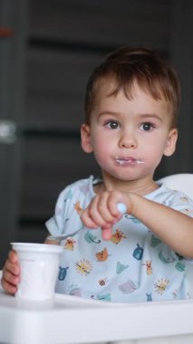 Yoğurttan hoşlanan sevimli erkek çocuk. Bebek mutfakta oturup plastik bardaktan süt ürünleri yiyor. Arkaplan bulanık. Dikey video