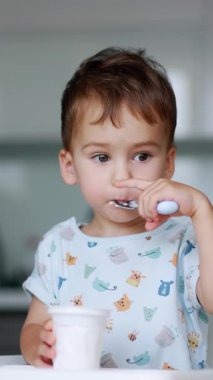 Düşünceli çocuk, elinde kaşıkla oturuyor. Bebek yoğurdunu yavaş yavaş yiyor. Arkaplan bulanık. Dikey video