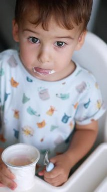 Küçük çocuk elinde yoğurtla beyaz sandalyede oturuyor. Bebek, biraz süt ürünleri alıp yemek için kaşık kullanıyor. Üst Manzara. Arkaplan bulanık. Dikey video