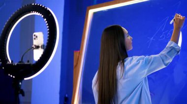 Koyu saçlı, beyaz saçlı, gömlekli bir kadın cam duvara beyaz kalemle yazı yazıyor. Kız telefona döner ve kamerayla konuşur..