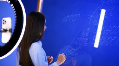 Esmer, kameraya konuşuyor ve cam duvardaki sayıları yuvarlak içine alıyor. Kurbağa veya akış videosu kaydediliyor.