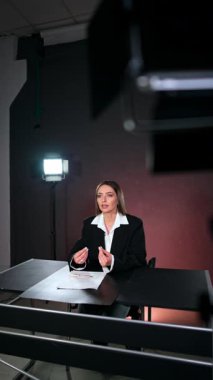 Koyu saçlı beyaz kadın stüdyoda masa başında oturuyor. Muhabir kamera içeriğiyle konuşuyor. Dikey video.