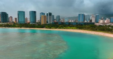 Pasifik Okyanusu 'nun turkuaz suyu üzerinden uçarak kumlu sahile yaklaşıyoruz. Gri bulutlu gökyüzünün altında yeşil kıyı ve güzel gökdelenler. Honolulu, Hawaii, ABD.