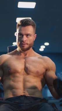 Genç üstsüz adam spor salonunda sıkı çalışıyor. Yakışıklı vücut geliştiren adam eğitimden sonra dinleniyor. Dikey video