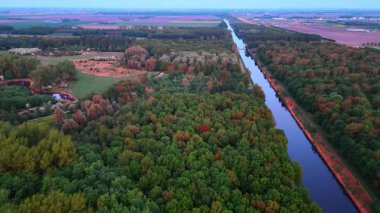 Kanaldan geçen şaşırtıcı yeşil yemyeşil orman. Tarımsal alanlar arka planda. Kırsal kesimde insansız hava aracı uçuşu.