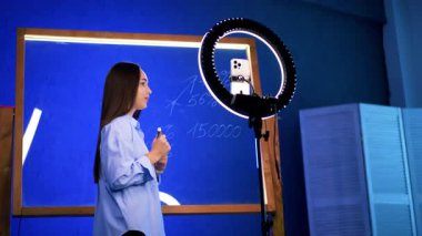 Modern sınıfta genç bir öğretmen. Genç bir kadın tebeşir tahtasının önünde duruyor, bir yandan dersi açıklarken diğer yandan da seyircileriyle iletişim kurmak için halka ışığını kullanıyor..