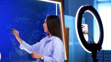 İnternette finans dersi veren genç bir kadın. Genç bir kadın, parlak bir stüdyoda online bir derste finansal kavramları anlatırken cam bir tahtaya yazı yazıyor..