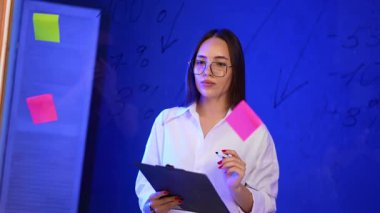 İş kadını fikirlerini sunuyor. Dürbünlü bir kadın, parlak, mavi ışıklı, yapışkan notlarla dolu bir çalışma alanında fikirleri tartışıyor..