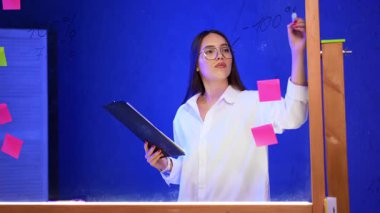 Genç profesyonel fikirleri paylaşıyor. Mavi bir duvarın önünde elinde renkli notlarla bir pano tutarken üzerine kalemler yazan bir kadın var..