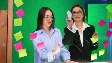 Stüdyoda beyin fırtınası yapan kadınlar. İki kadın cam bir masada iş birliği yapıyor, yaratıcı bir alanda canlı renklerle çevrili yapışkan notlar yazıyorlar..