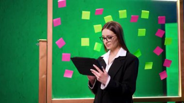 Ofiste fikirleri paylaşan bir kadın. Siyah takım elbiseli bir kadın yapışkan notlarla dolu renkli bir beyin fırtınası ortamında tabletten okuyor..