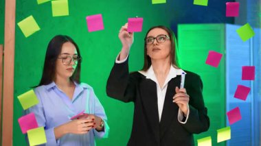 Ofiste beyin fırtınası yapan kadınlar. İki kadın beyin fırtınası sırasında cam bir tahtadaki yapışkan notları kullanarak yaratıcı bir şekilde iş birliği yapıyorlar..