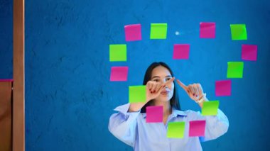 Beyin fırtınası yapış yapış. Genç bir kadın mavi bir duvara renkli yapışkan notlar yerleştirirken gülümsüyor ve beyin fırtınası yapıyor..