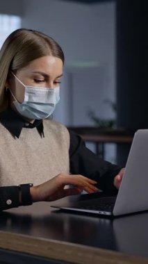 Dizüstü bilgisayarla çalışan maskeli bir iş kadını. Karantina serbest çalışanı işyerinde çalışıyor. Dikey video