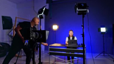 Tripod 'daki kamera stüdyodaki bir kadını kaydediyor. Kadın bilgisayarında çalışıyor. Arkaplanda bulanık duruyor. Erkek fotoğrafçı masaya gelip bir şey koyuyor..