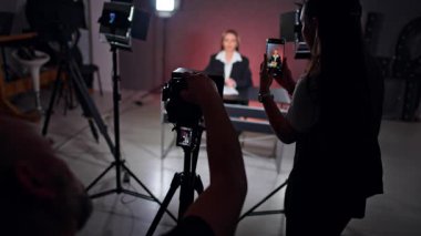 Siyah ceketli kadın profesyonel kamera ve akıllı telefondan çekiliyor. Stüdyoda günlük içeriği oluşturma. TV program kaydı.