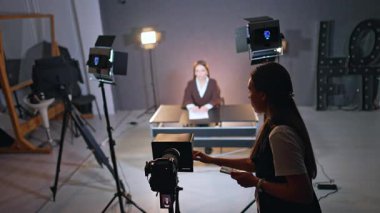 Elinde telefon olan genç, uzun saçlı kadın tripodun üzerinde kameraya dokunuyor. Arka plandaki bayan bulanık arkaplanda masasında oturuyor. Stüdyoda TV veya blog görüntüleri.