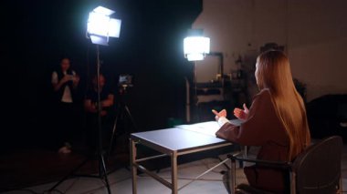 Esmer, uzun saçlı bir kadının stüdyoda video kaydının arka görüntüsü. Erkek kameraman ve bayan asistan sahne arkasında çalışıyor..