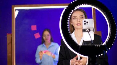 Bayan telefon ve halka lambasının önünde konuşuyor. Kadın video kaydediyor ya da blog için yayın yapıyor. Arka plandaki kız cam duvara etiket yapıştırıyor..