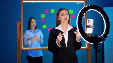 Siyah ceketli kadın elinde kamera ve halka ışığıyla tripodun önünde duruyor. Bayan konuşuyor ve ellerini sallıyor. Bir kız gelir ve kamerayla konuşur. Günlük konsepti.