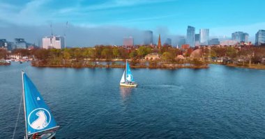 Mavi yelkenli tekneler güneşli bir günde Charles Nehri 'nden geçer. Boston 'un silueti arka planda kalın bir sis tabakasıyla kaplıdır..