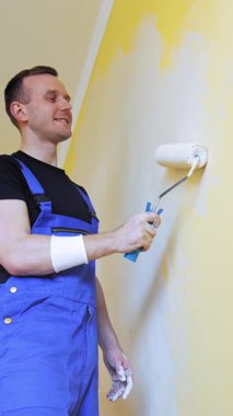 Evdeki duvarları boyayan adam. İş yerindeki yakışıklı adam tulumuyla duvarı silindirle boyuyor.
