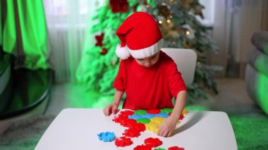 Çocuk bulmacayı ağaçtan yapar. Kırmızı tişörtlü ve Noel Baba şapkalı bir çocuk rengarenk yapboz parçaları toplarken, yanında süslü bir Noel ağacı parlıyor..