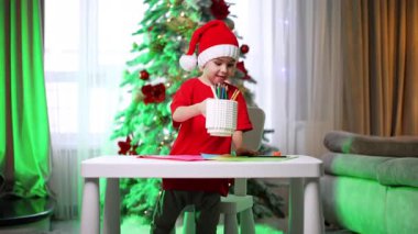 Çocuk evde el işi yapıyor. Şenlik şapkası takan neşeli bir çocuk süslü bir Noel ağacının yanındaki masada el işi yapıyor..