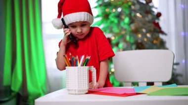 Noel Baba şapkalı çocuk şenlik çağrısı yapıyor. Noel Baba şapkası takan bir çocuk telefonda konuşurken etrafı renkli kağıtlarla çevrili..