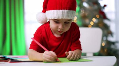 Çocuk sevinçle tatil zanaatı yapıyor. Genç bir çocuk, sezon için dekore edilmiş bir masada tatil sanatı çizerken şenlikli bir şapka takıyor..
