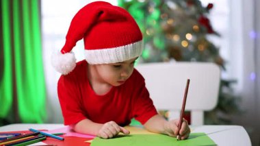 Çocuk bayram kutlaması sırasında resim çizer. Kırmızı tişörtlü ve Noel Baba şapkalı bir çocuk süslü bir Noel ağacının yanındaki masada renkli sanatlar yaratıyor..